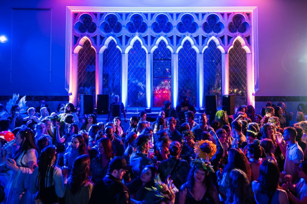 A lively crowd dancing in front of tall arched windows.