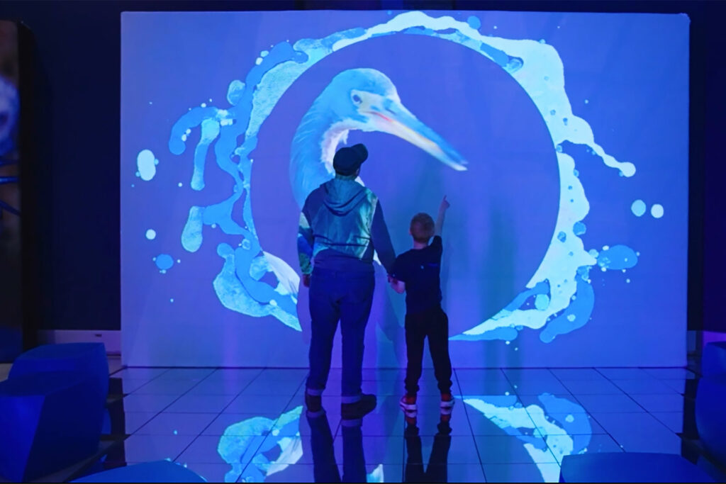 A man and a boy, facing away, stand in front of a projection of clouds.
